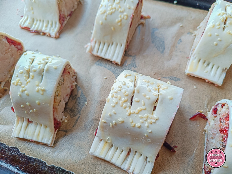 Unbaked gluten-free sausage rolls brushed with egg and sprinkled with sesame seeds, laid out on parchment.